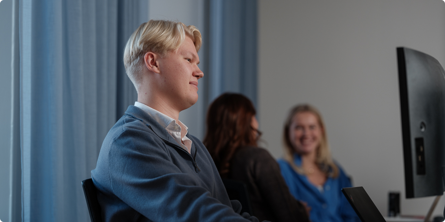 kontor med en person som sitter närmast och kollar in i skärmen med två andra personer i bakgrunden som pratar och ler.