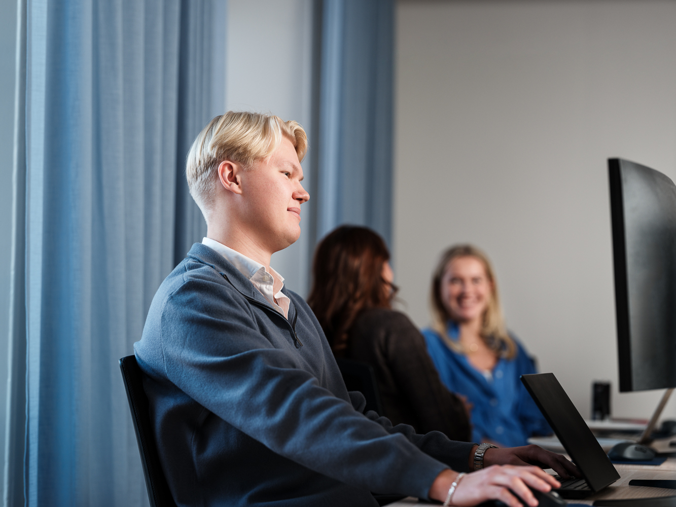 Kille som sitter och jobbar framför dator vid skrivbord, två personer sitter i bakgrunden och pratar