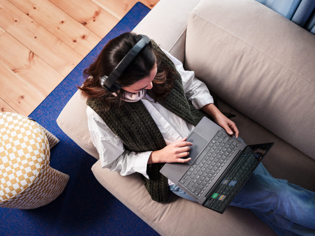 Tjej som sitter och jobbar med laptop och hörlurar i en soffa.